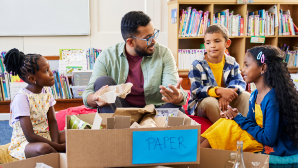 An adult sits between boxes labeled for different types of recyclables, teaching children.
